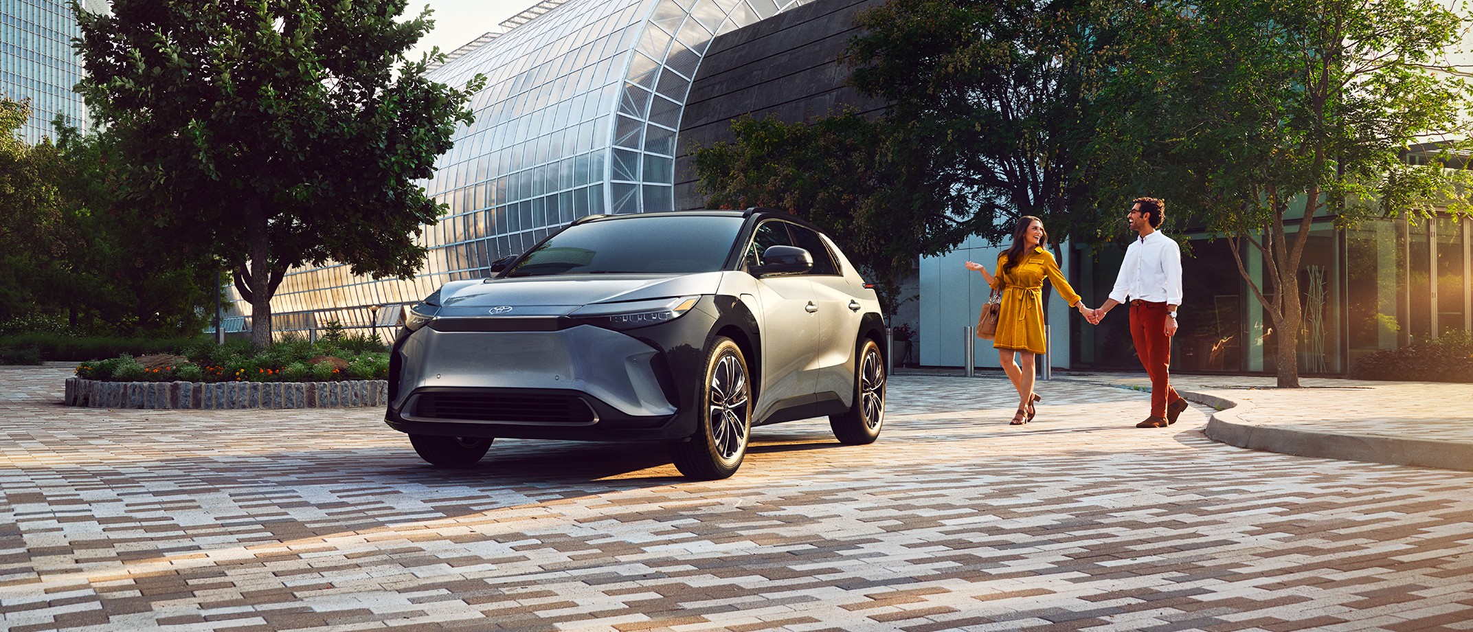 Image of a couple in bright clothing walking to their electrified vehicle with city building in background.