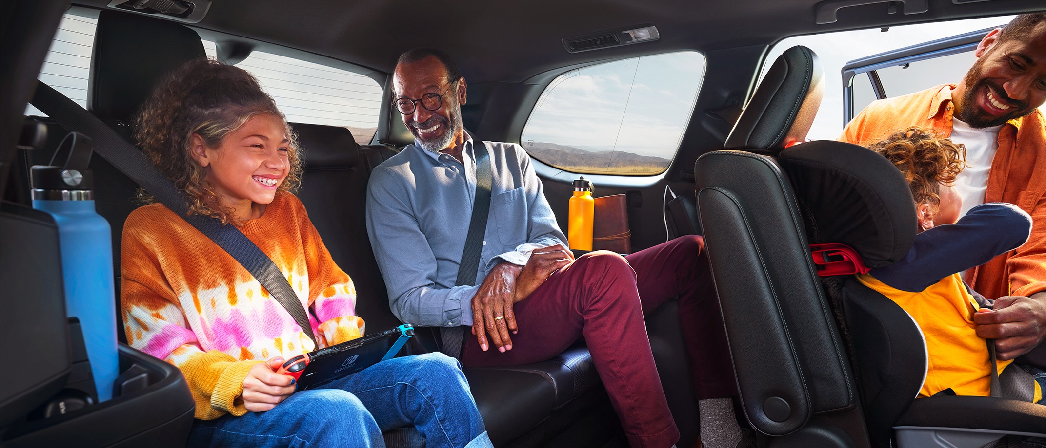 Image of a family wearing clothing in bright, saturated color sitting inside their electrified vehicle. A grandfather sits smiling at his teengage grandaughter next to him. In the front, a father helps his toddler into a car seat.