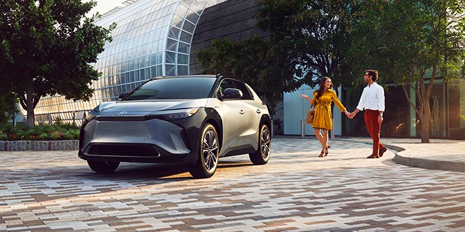 Image of a couple in bright clothing walking to their electrified vehicle with city building in background.