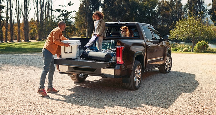 A Toyota Tundra parked in front of a cabin. A dad grabs a cooler from the trunk with his daughter and dog.