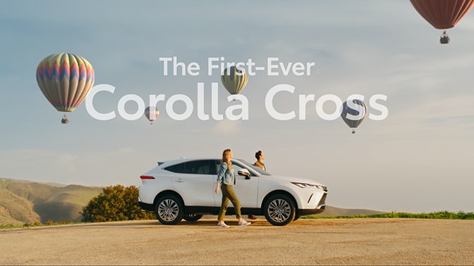 Image of couple getting out of vehicle with hot-air balloons in the sky. At the top of the image, “The First Ever Corolla Cross” is written in white text that is starting to fade out.