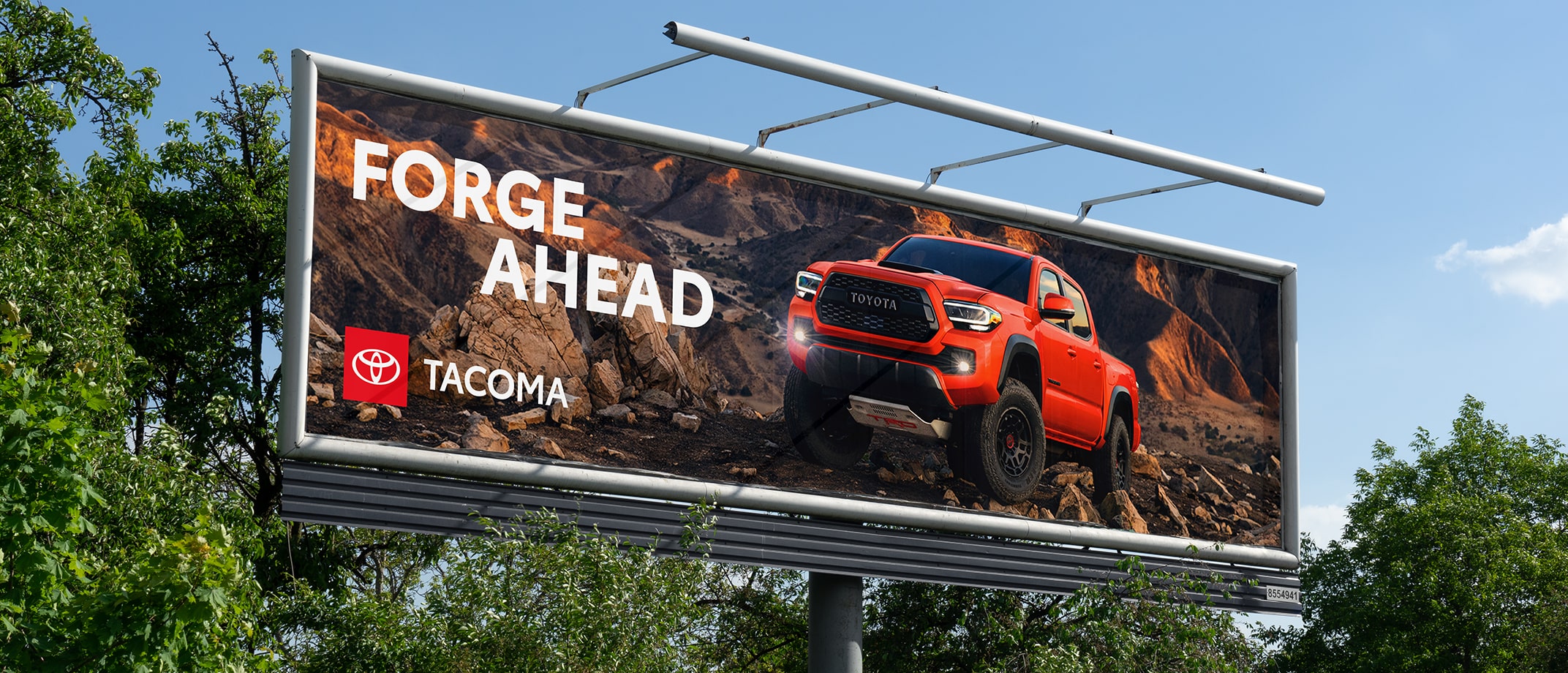An out-of-home billboard with a Tacoma logo shows a Tacoma parked on the top of a rocky mountain, the headline reading “Forge Ahead.”