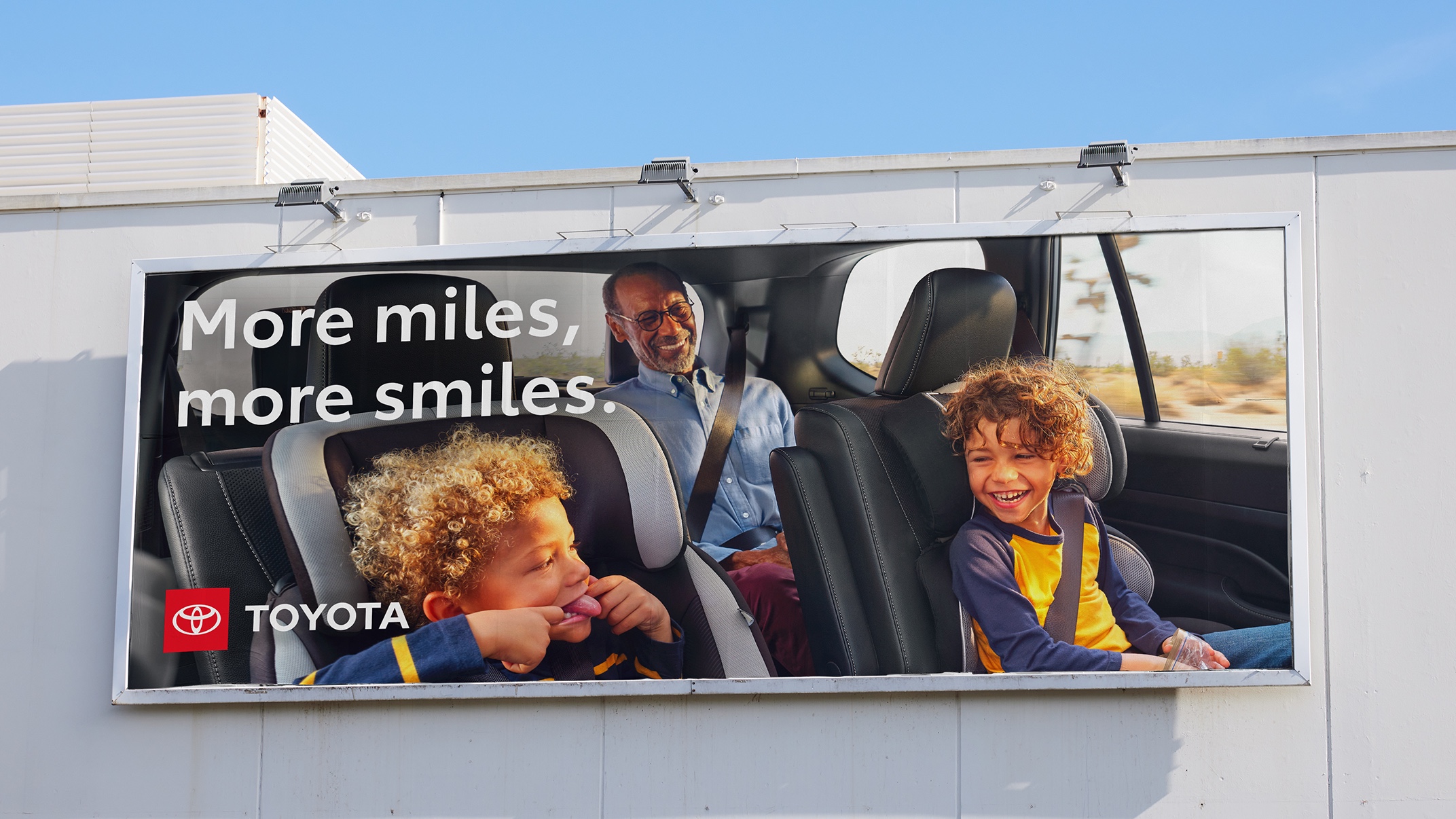 An out-of-home billboard with the Toyota logo shows a family inside their Grand Highlander. The headline reads “More miles, more smiles.”