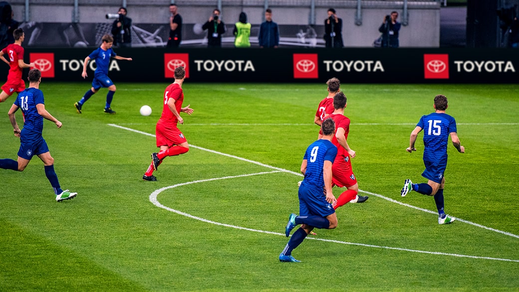 Image of soccer teams on a field with the Toyota logo displayed on the perimeter wall against a black background.