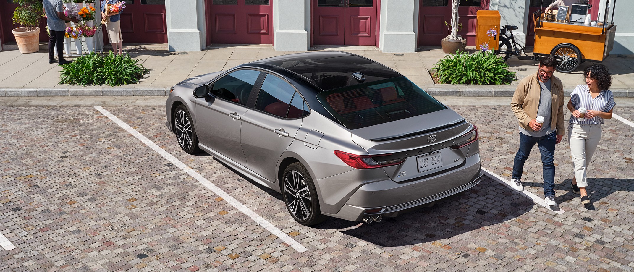 A man and woman holding cups walking past car parked on cobblestone parking spot.