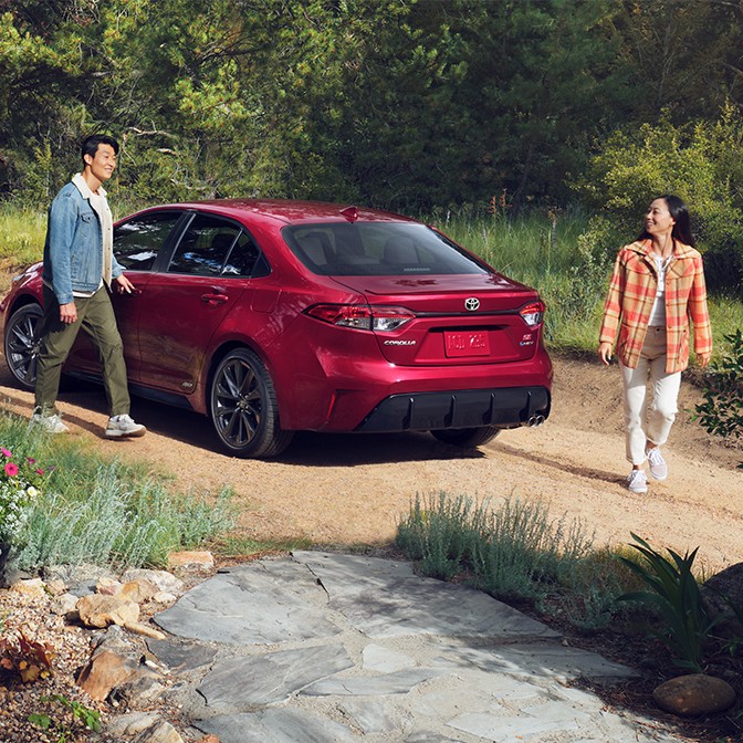 A woman and man standing outside of a red Toyota parked on a dirt road.