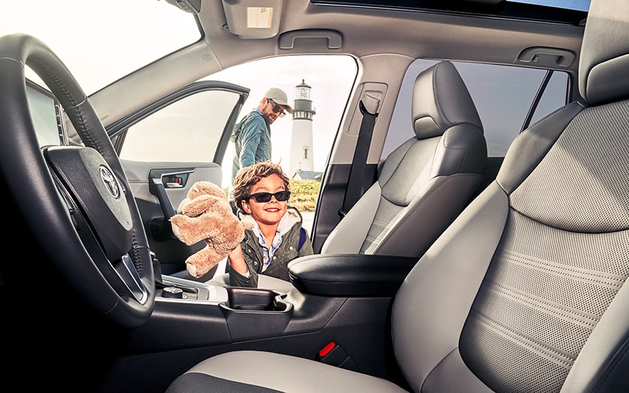 A young child holding a stuffed animal gets into a vehicle with their dad in the background.