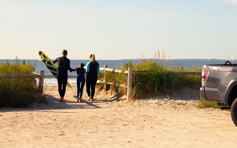 A family in surf gear with a surfboard. The mom and dad assist their child as they walk toward the ocean. The tail end of a Tundra can be seen in the foreground.