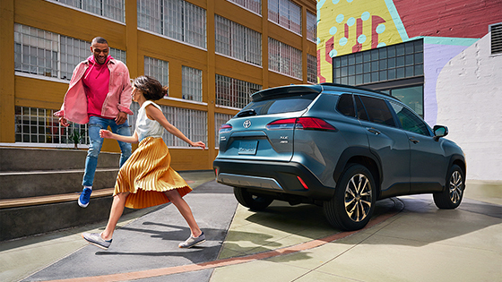 Man and woman in bright clothing walking behind a Toyota.