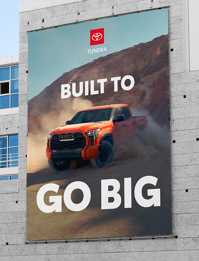 An out-of-home billboard on the side of a building shows a Toyota Tundra kicking up dust. The headline is in all-caps reading “BUILT TO GO BIG,” with “GO BIG” in a larger, bolder font weight.
