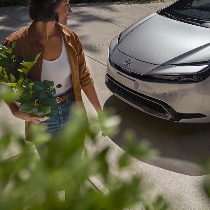 A photograph of a woman holding a plant looking back at her car.