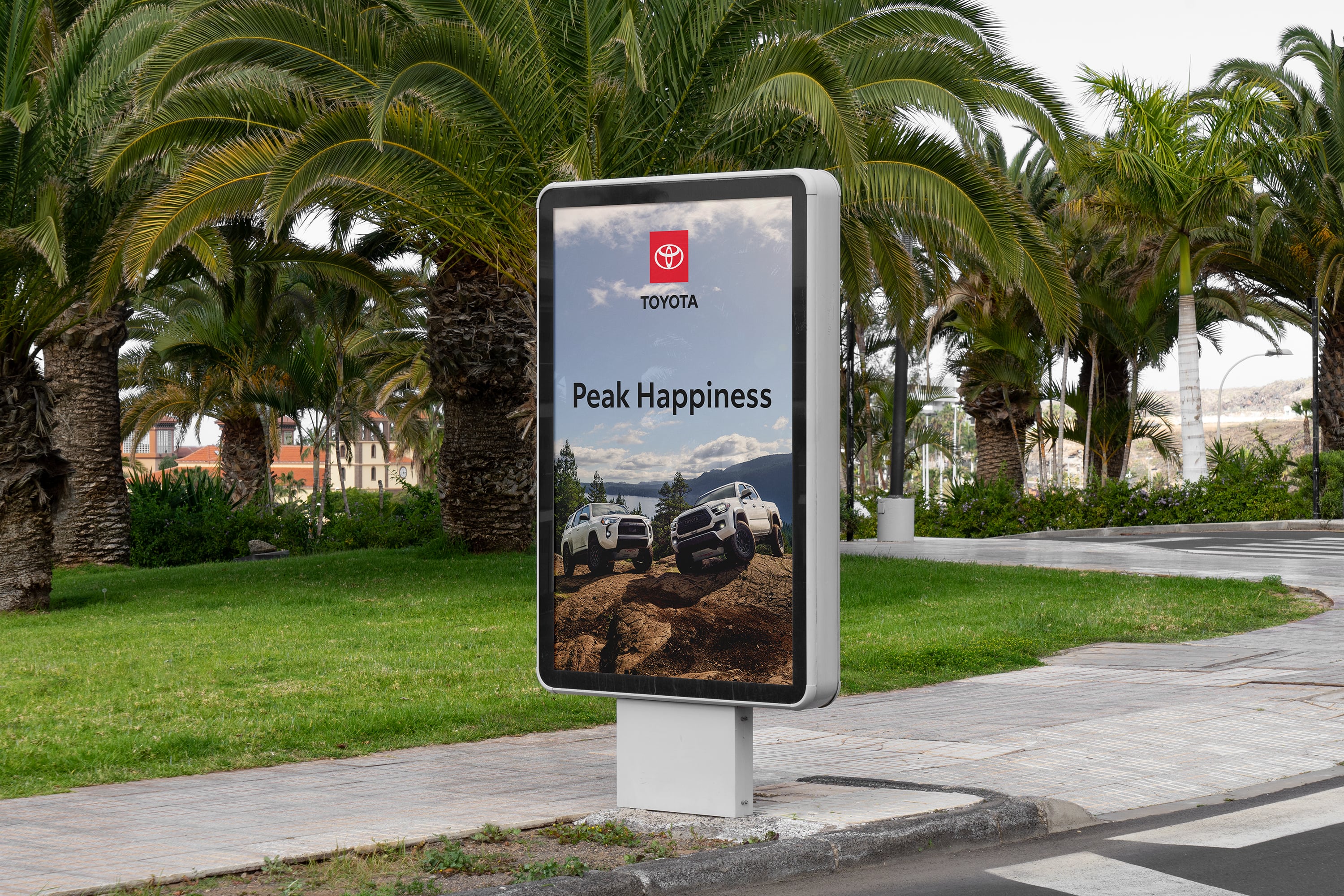 A digital out-of-home sign in a park displays an add with the Toyota logo, headline and two Toyota trucks atop a mountain.