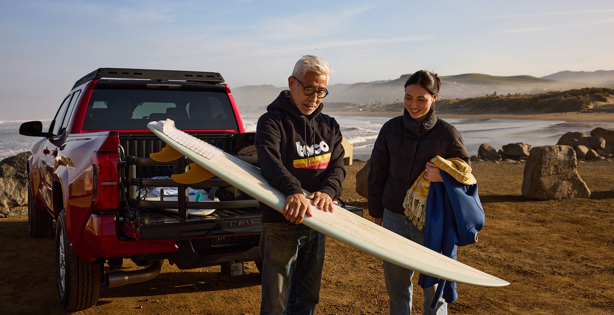  A man holding a surfboard with a woman standing next to him with a red truck behind them and a backdrop of the ocean and mountains.
