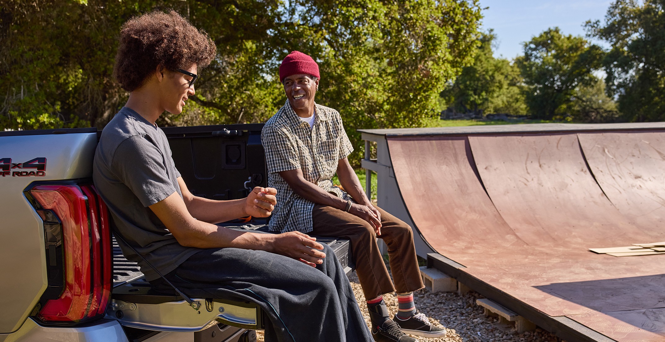 Two men sitting on the trunk of a truck with a skateboard ramp next to them.
