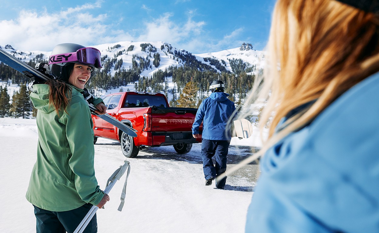 In a snowy mountain landscape, a woman wears a winter jacket and ski mask. She has ski gear and smiles at a girl in the foreground. In the background, a man walks towards a red Toyota truck