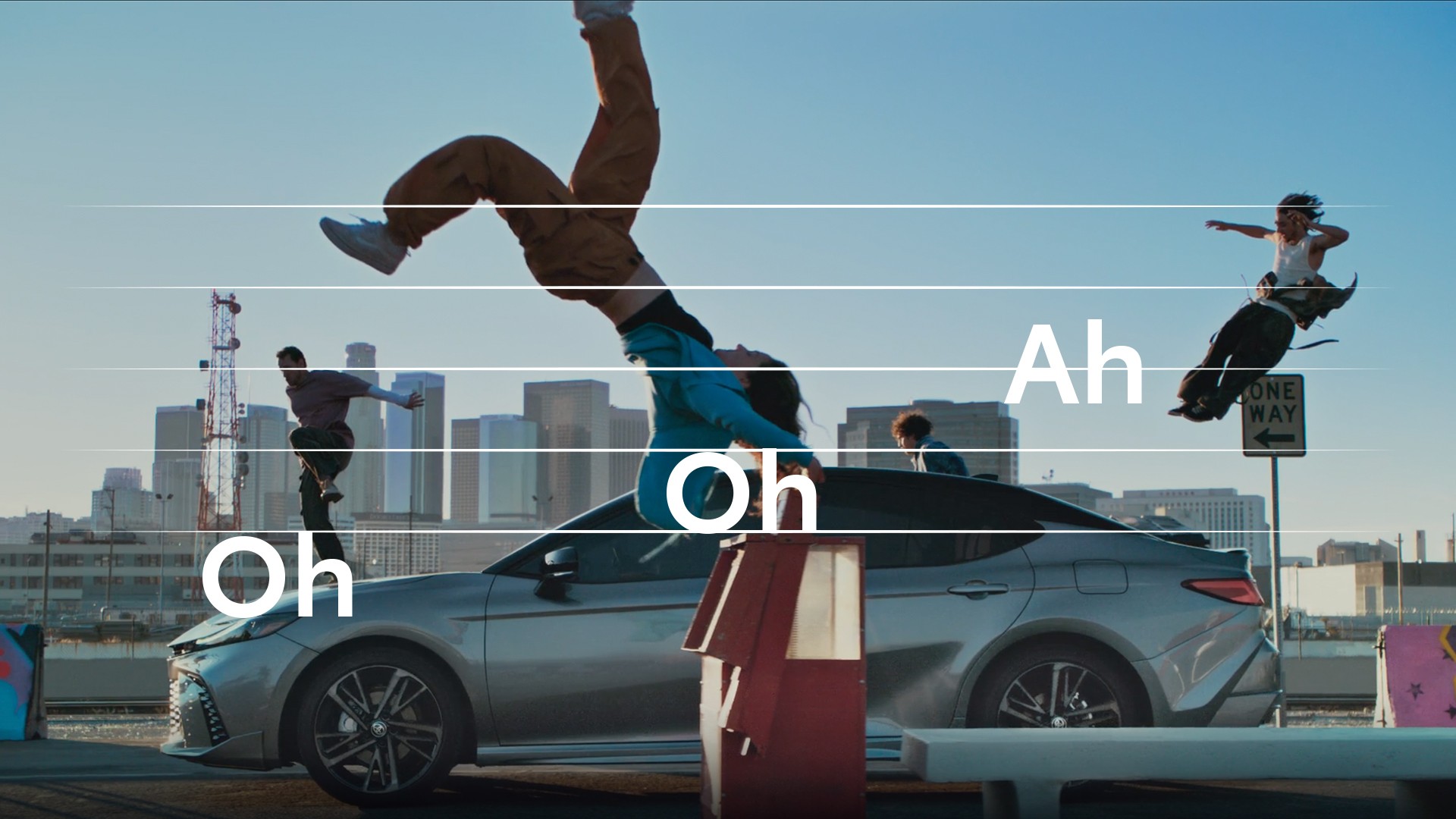 An image of people dancing outside of a vehicle to music with a super of the words “Oh Oh Ah” displayed like musical notes.
