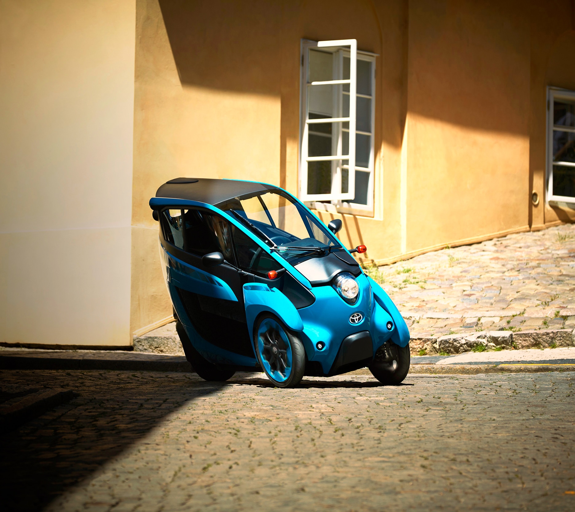 Blue Toyota i-ROAD Personal Mobility Vehicle (PMV) parked on a cobblestone street.