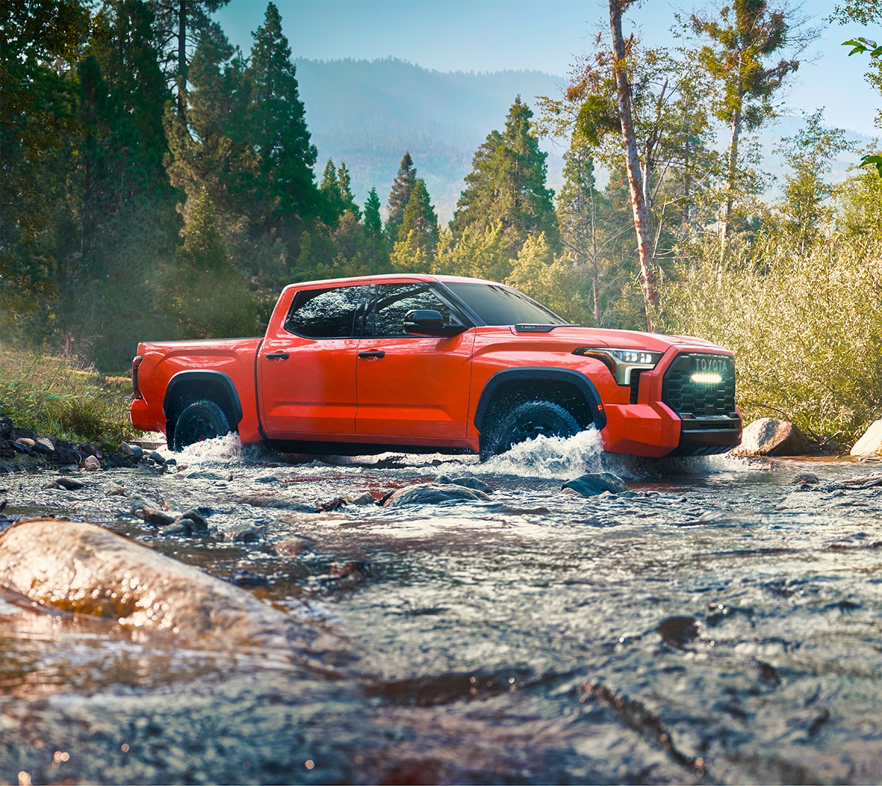 A red Toyota truck crosses a creek with trees and mountains in the background.