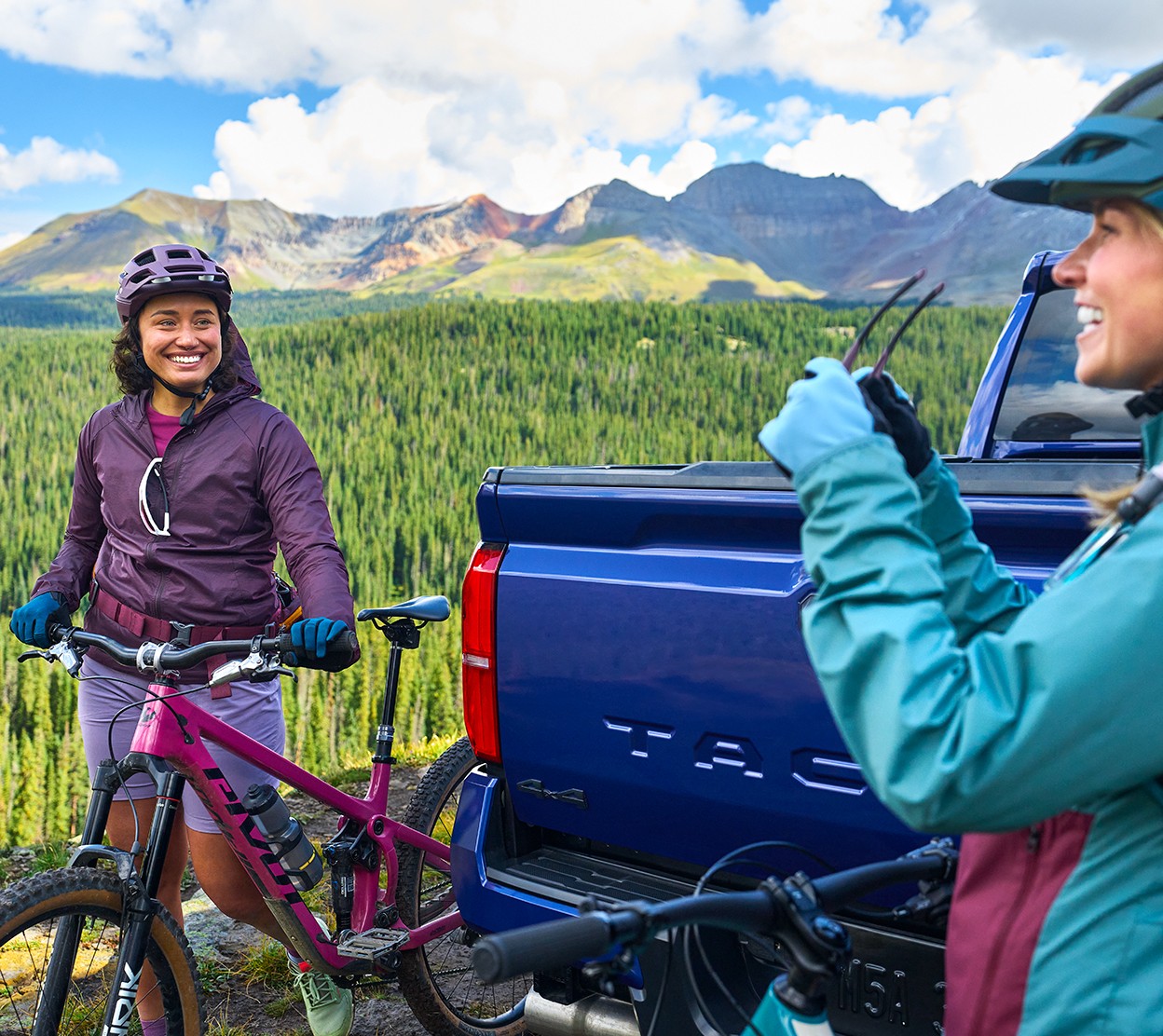 In a scenic landscape, two woman with bikes wearing biking gear hang out together at the back of their Toyota truck preparing for a mountain biking adventure.