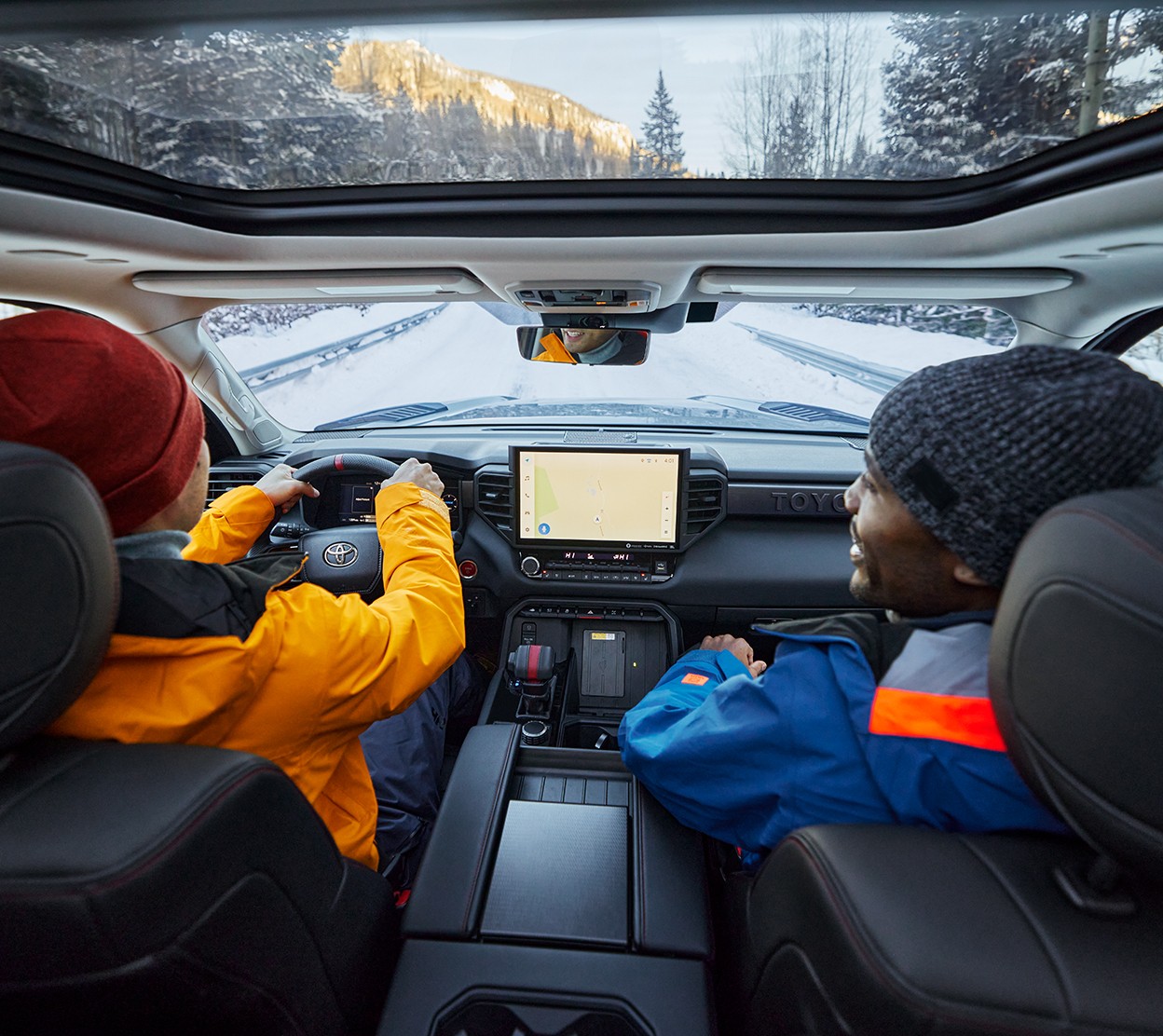 Inside a Toyota vehicle, a couple wearing bright clothing drives down a snowy road. The man smiles towards the driver and through the windshield and moonroof we see a snowy forest.