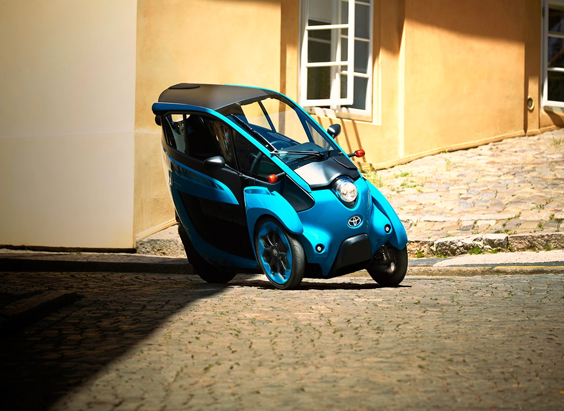 Blue Toyota i-ROAD Personal Mobility Vehicle (PMV) parked on a cobblestone street.