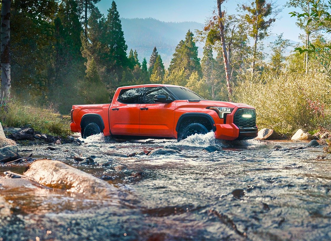A red Toyota truck crosses a creek with trees and mountains in the background.