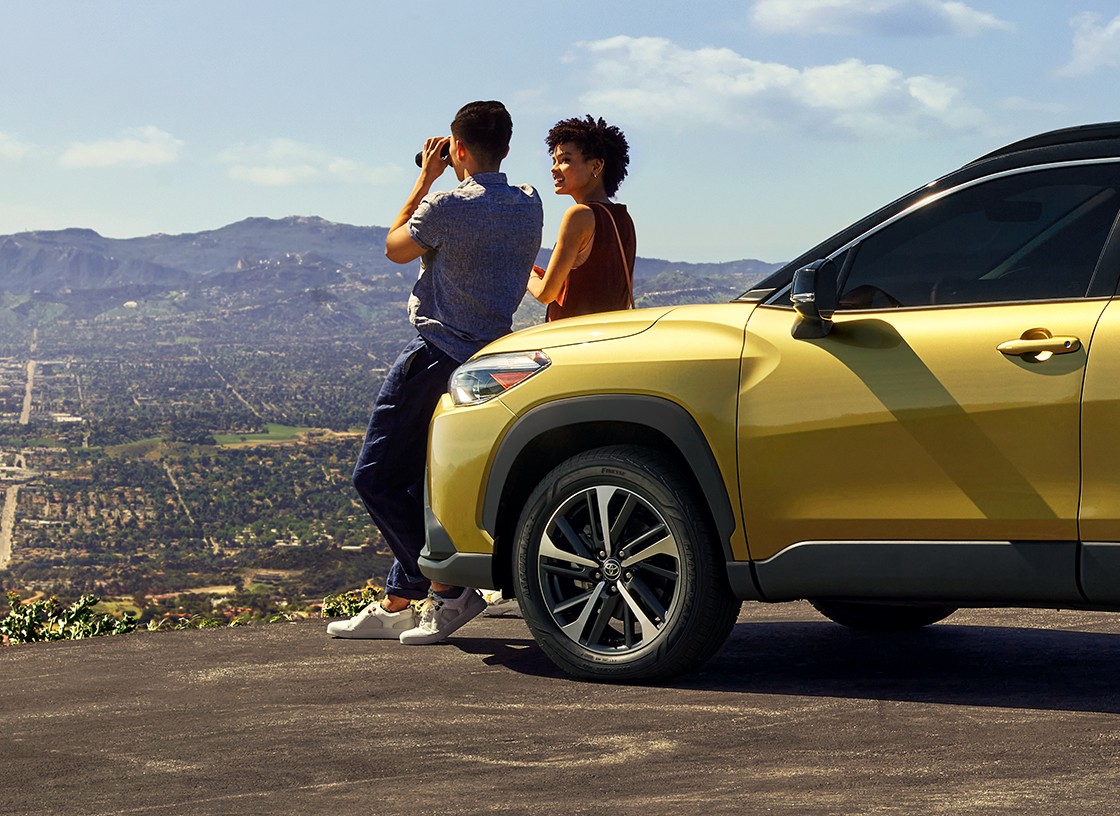 At a scenic viewpoint, a couple leans against the hood of a bright yellow vehicle. The man holds binoculars as they gaze out at the mountains.