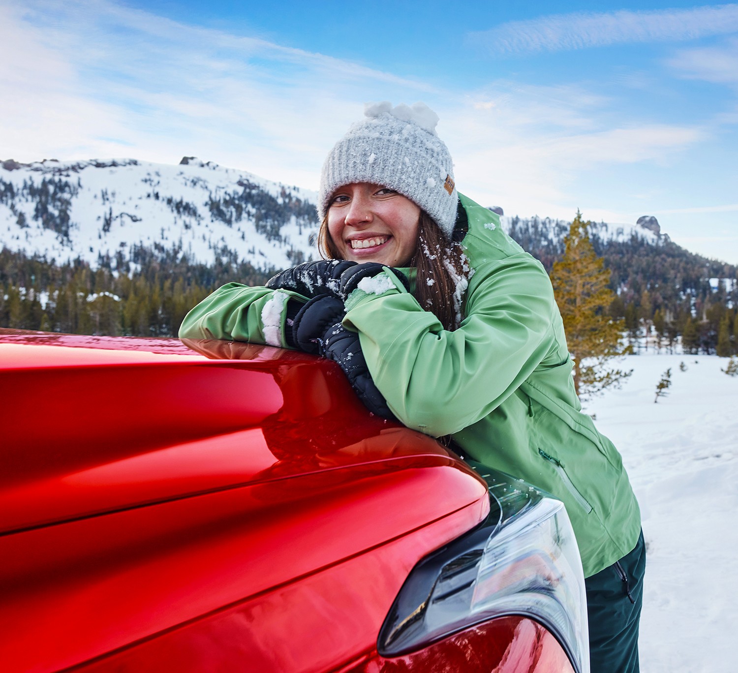 In a scenic winter landscape, a woman leans against the hood of a Toyota vehicle with her head propped on her hands smiling at the camera.