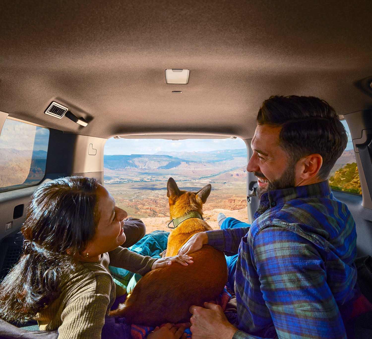 A couple lounge in the back of their Toyota truck laughing with each other while their dog looks out through the open tailgate at a scenic landscape.