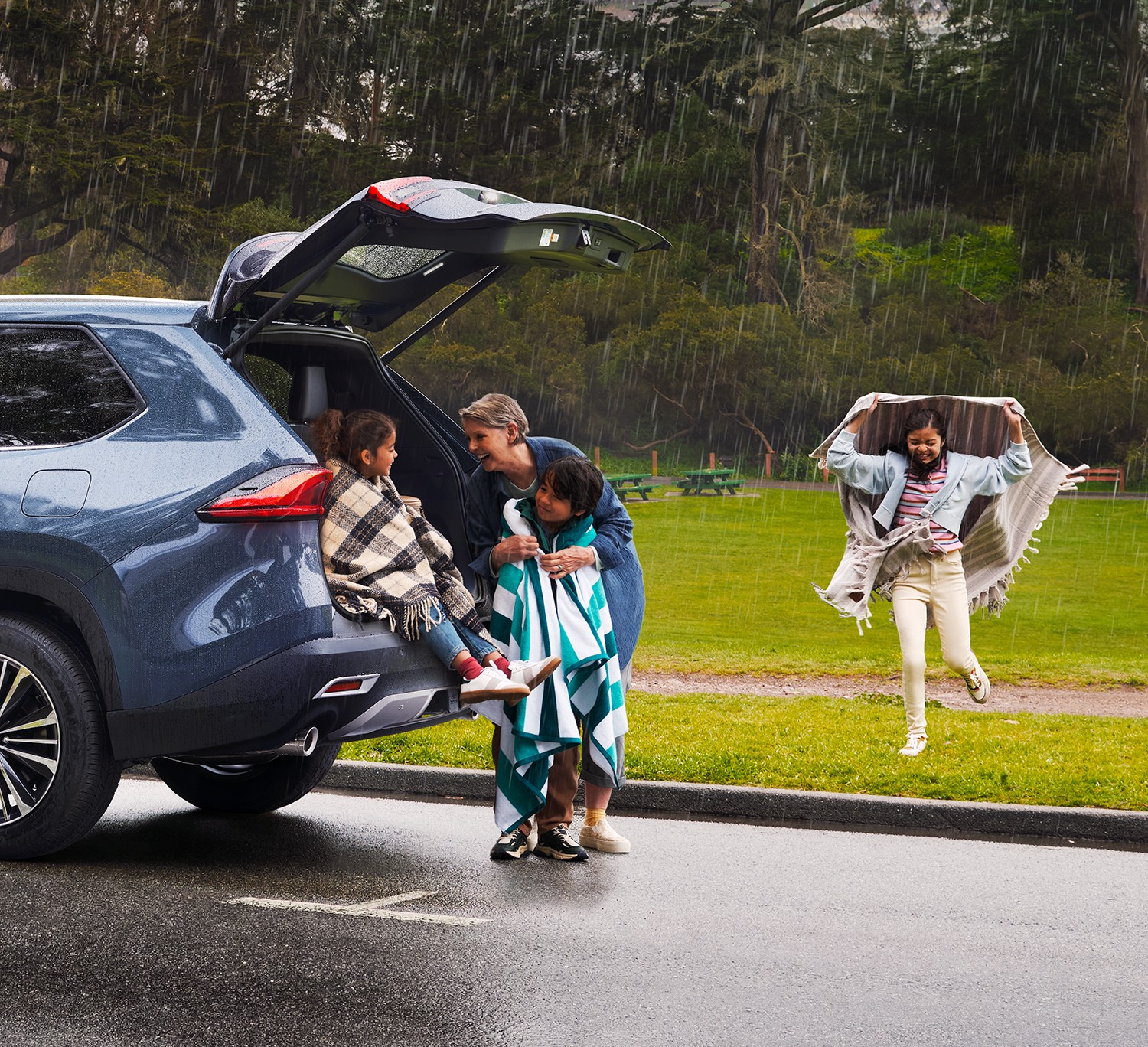 On a rainy day at the park, two kids sit at the back of their Toyota while their mom laughs and dries them off with towels. In the background, a girl runs towards the car holding a towel over her head to protect her from the rain.