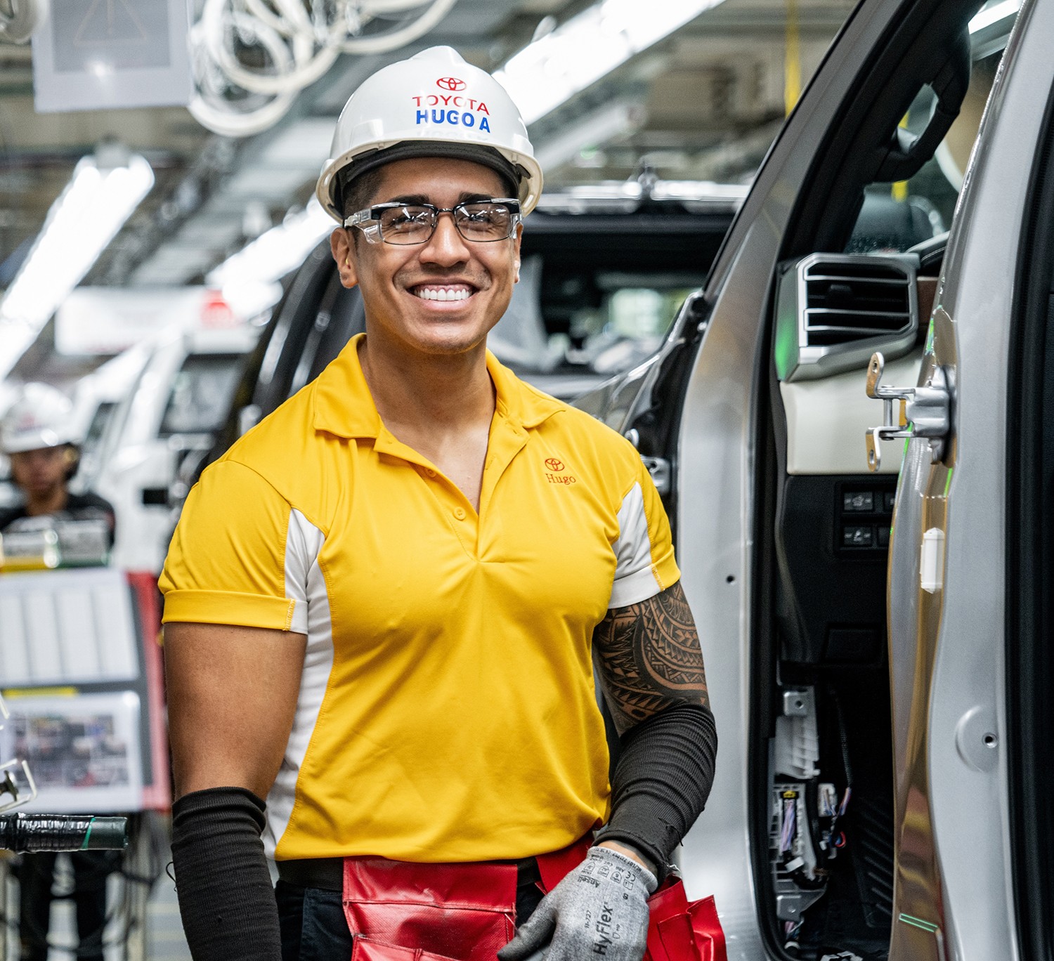 A factory worker in a yellow shirt and helmet stands next to a vehicle on the assembly line and smiles at the camera.