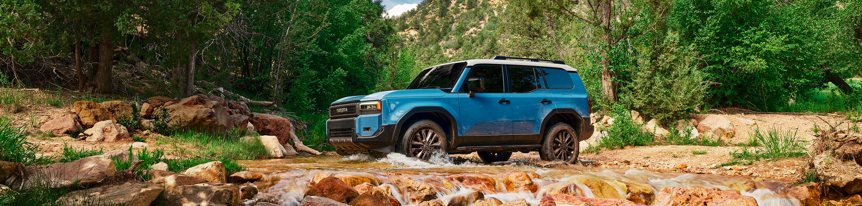 A blue Toyota Land Cruiser with a white roof crosses a creek in a forest valley.