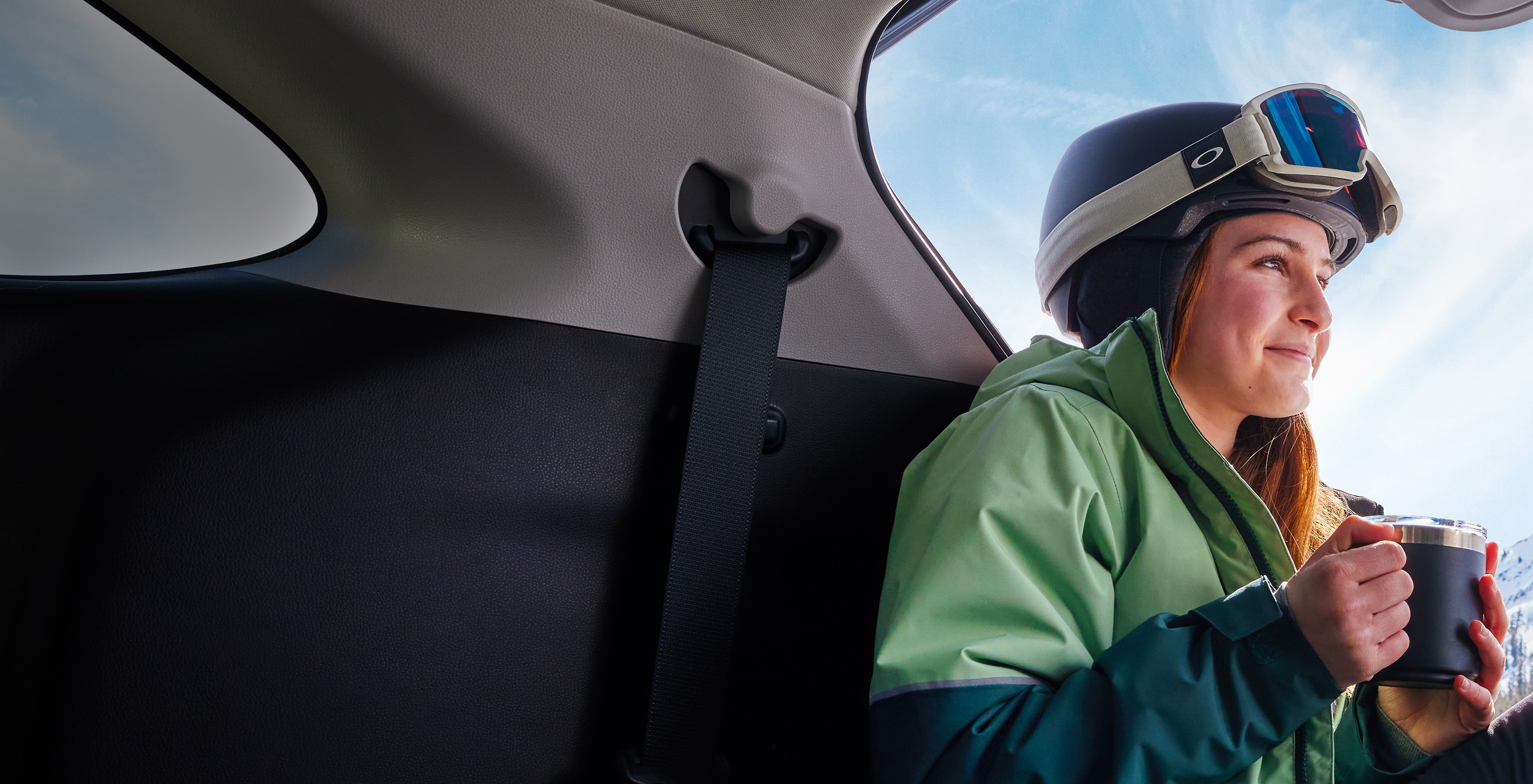 A woman wearing snowboarding gear sits inside a Toyota vehicle. She’s holding a mug of coffee and looks out the window smiling.